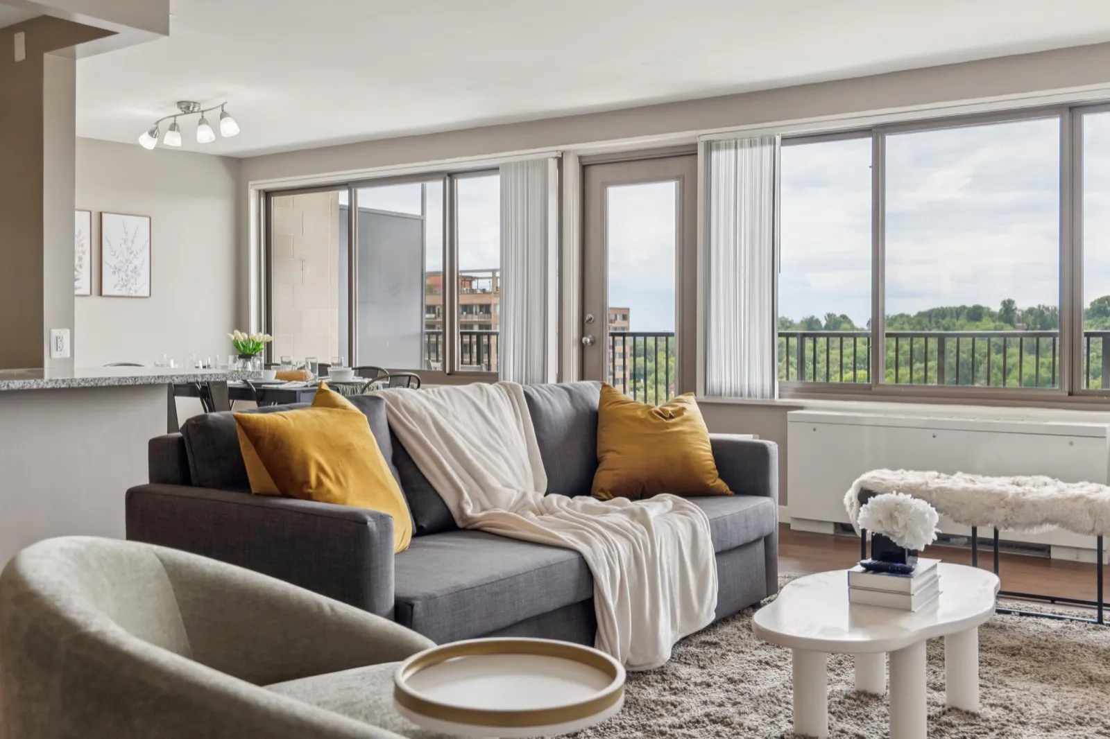 Bright furnished living room with a gray sectional, gold accent pillows, and wraparound windows over a tree-lined view in Crystal City, Arlington VA